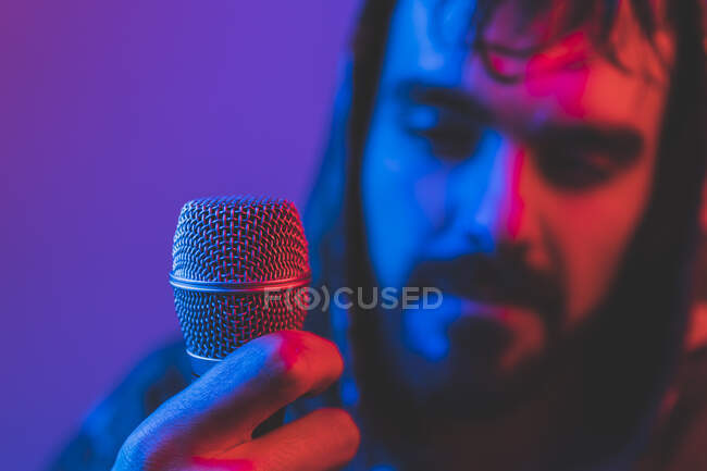 Maschio adulto barbuto cantante in giacca con cappuccio e microfono durante concerto rock in illuminazione al neon — Foto stock