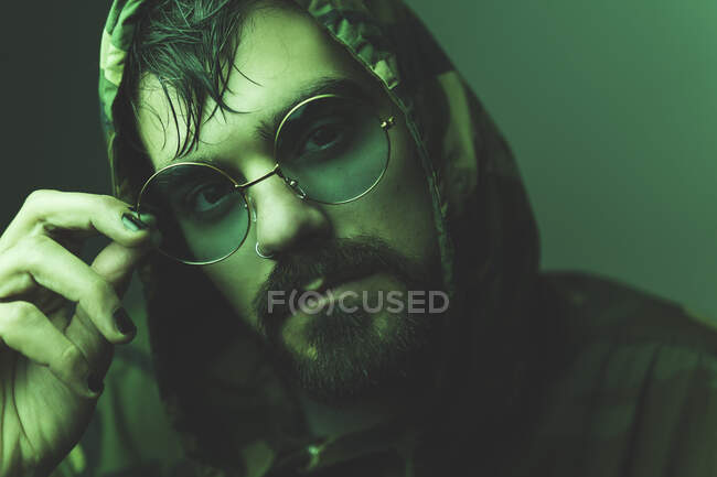 Pensive bearded hipster male in sunglasses and camouflage hooded jacket standing and touching glasses in studio with green illumination — Stock Photo