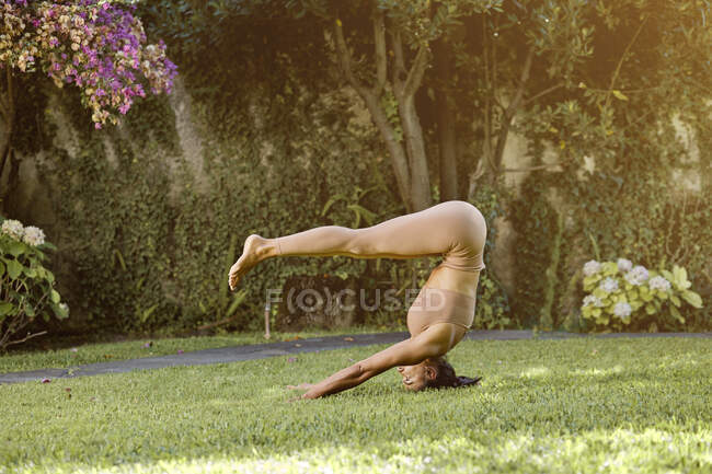 Vue latérale de heureuse femme flexible dans les vêtements de sport démontrant Adho Mukha Vrksasana pose avec les jambes plier tout en pratiquant le yoga sur tapis et détourner les yeux — Photo de stock
