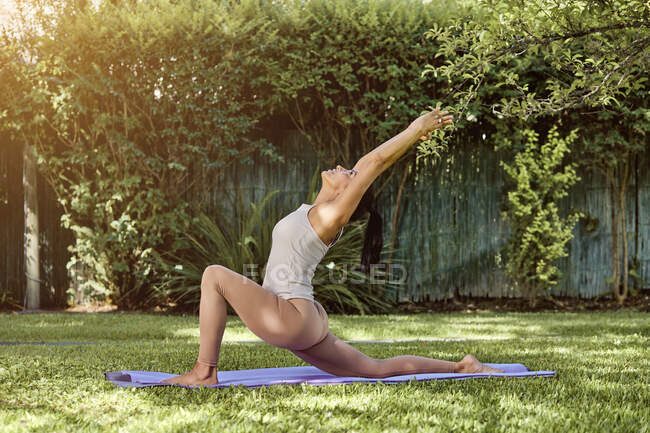 Vue latérale de la femme flexible dans les vêtements de sport démontrant la pose Anjaneyasana tout en pratiquant le yoga sur tapis et en levant les yeux — Photo de stock