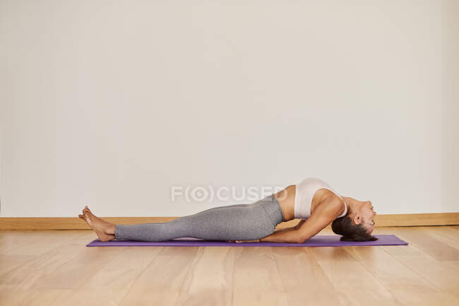 Vista laterale della femmina a piedi nudi in vestiti sportivi che praticano yoga sul pavimento contro il muro bianco nell'edificio — Foto stock
