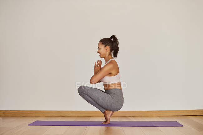 Side view of smiling adult female in sportswear balancing on yoga mat with namaste hands and looking forward — Stock Photo