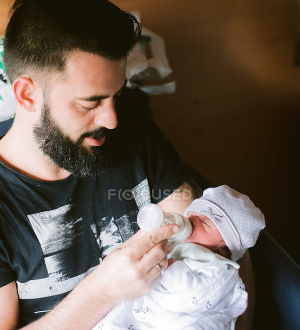 High angle of loving adult bearded dad with milk bottle feeding tiny newborn kid and enjoying first moments of fatherhood at home — Stock Photo