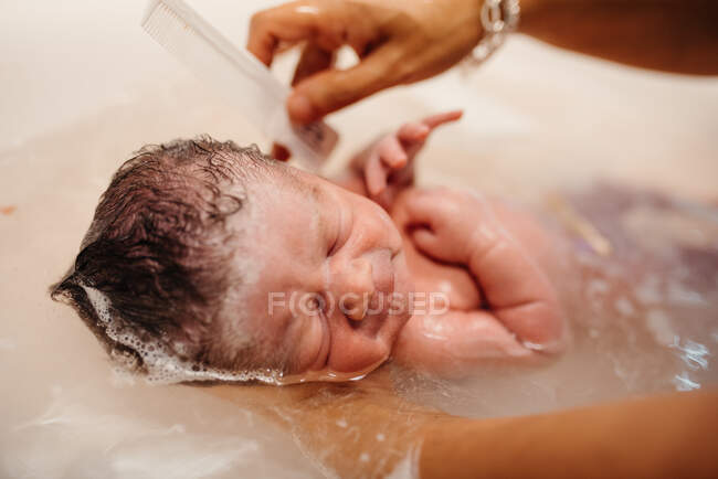 High angle of cute newborn child held by anonymous mother with comb getting bath in sink — Stock Photo