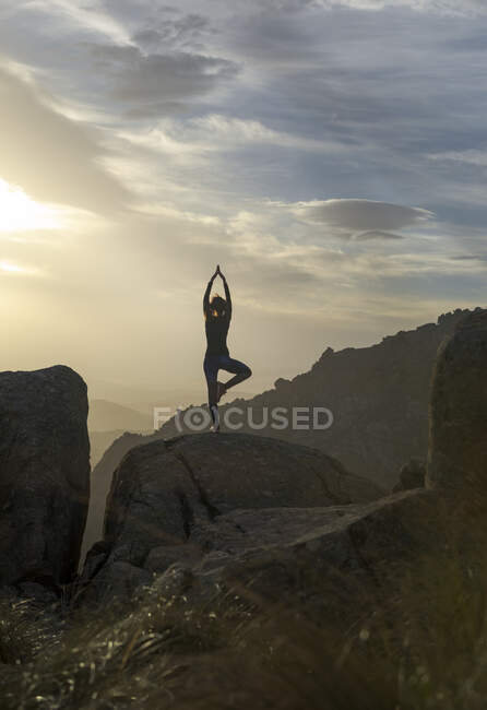 Vista posterior de la hembra irreconocible de pie en la cima de la montaña rocosa y haciendo la pose Árbol con los brazos hacia arriba durante la puesta del sol - foto de stock