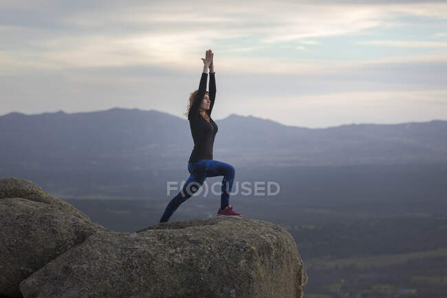 Side view of unrecognizable female standing on top of rocky mountain and doing Warrior one pose during sunset — Stock Photo