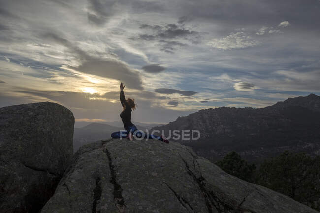 Vue latérale de la femelle méconnaissable debout sur le sommet de la montagne rocheuse et faisant Pigeon Pose pendant le coucher du soleil — Photo de stock