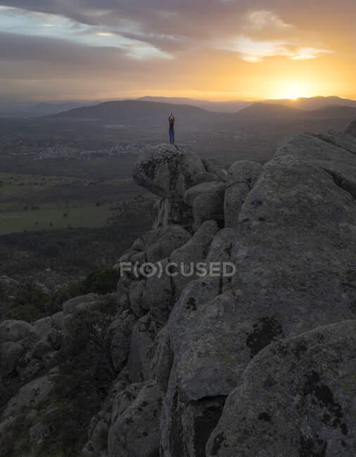 Visão traseira da fêmea irreconhecível em pé no topo da montanha rochosa e fazendo Árvore com braços para cima pose durante o pôr do sol — Fotografia de Stock