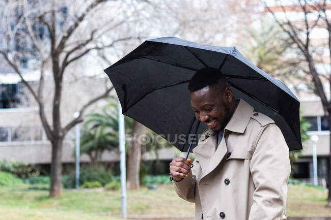 Trendiger erwachsener afroamerikanischer Mann in stylischem Trenchcoat mit schwarzem Regenschirm lächelt strahlend in die Kamera, während er an einem Frühlingstag auf der Straße steht — Stockfoto