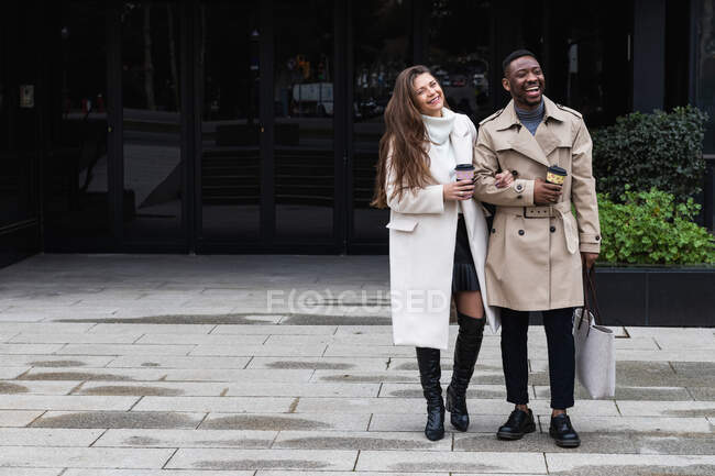 Ganzkörper fröhliches erwachsenes multiethnisches Paar in trendigen Mänteln mit Tassen Kaffee zum Mitnehmen auf gepflasterter Stadtstraße — Stockfoto