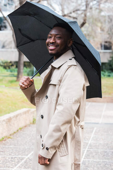Trendy adult African American male in stylish trench coat with black umbrella smiling brightly and looking at camera while standing on urban street in spring day — Stock Photo