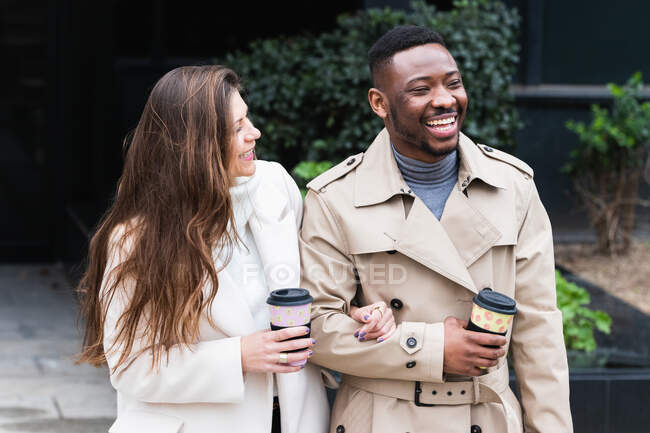 Fröhliches erwachsenes multiethnisches Paar in trendigen Mänteln mit Tassen Kaffee zum Mitnehmen auf gepflasterter Stadtstraße — Stockfoto