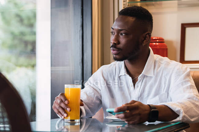 Serious adult African American sitting at table using smartphone while sipping an orange juice in a coffee shop — Stock Photo