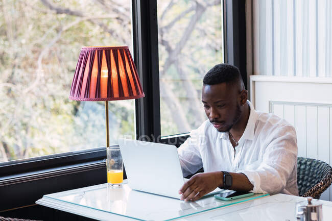 Concentrated African American male remote specialist using laptop while sitting at table with glass of juice in cozy cafe — Stock Photo