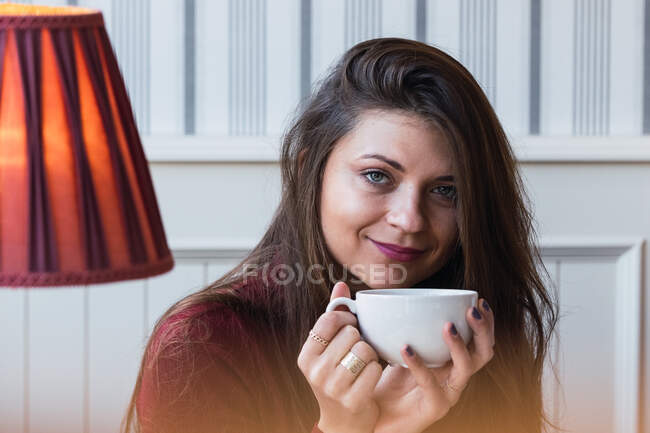 Donna adulta sognante con tazza di caffè caldo nelle mani guardando la fotocamera e sorridendo mentre si gode il tempo libero nel caffè — Foto stock