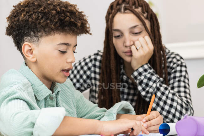 Бічний погляд на урожай чорний школяр робить нотатки в зошиті і вчиться за столом у будинку, а його сестра виправляє його — стокове фото