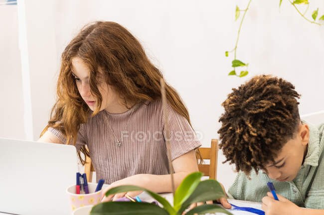Aufmerksame Schülerin benutzt Netbook in der Nähe von schwarzem Jungen, der auf Papier schreibt, während er Hausaufgaben macht — Stockfoto
