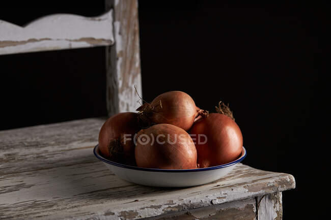 Cuenco con cebollas enteras sin pelar colocado en una silla de madera de mala calidad - foto de stock