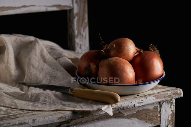 Чаша с целым неочищенным луком помещена рядом с льняной салфеткой и ножом на потрепанном деревянном стуле — стоковое фото