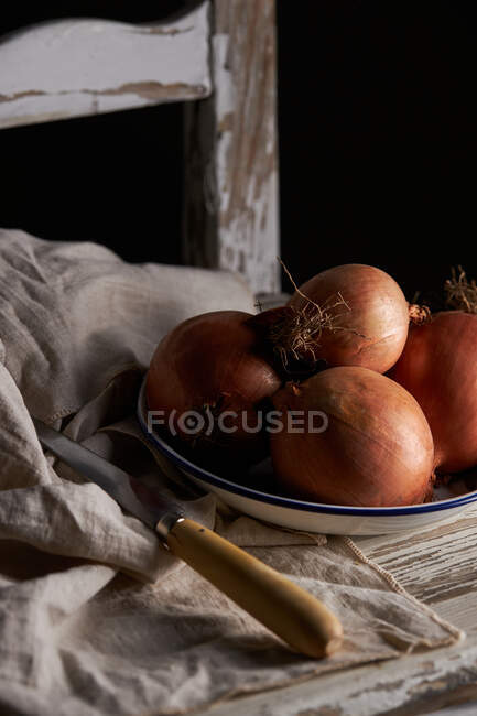 Cuenco con cebollas enteras sin pelar colocadas cerca de servilleta de lino y cuchillo en una silla de madera de mala calidad - foto de stock