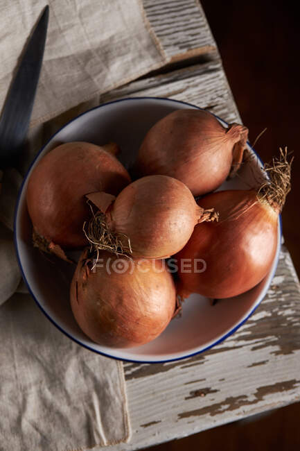 Top view of bowl with whole unpeeled onions placed near linen napkin and knife on shabby wooden table — Stock Photo