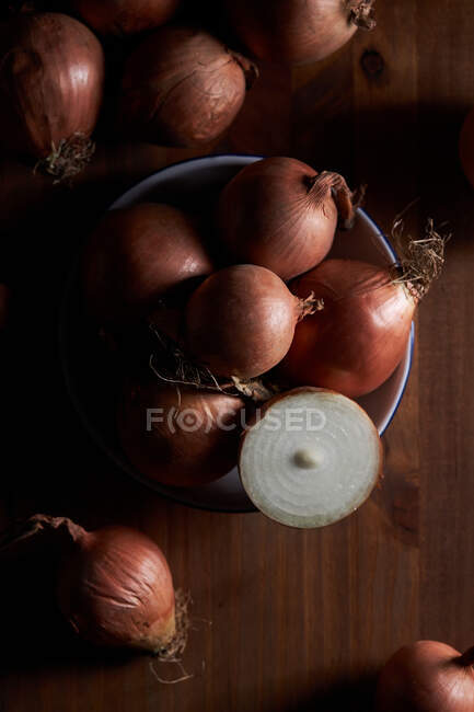 Nahaufnahme halbiert und ganze ungeschälte Zwiebeln in Schüssel auf schäbigem Tisch auf schwarzem Hintergrund platziert — Stockfoto