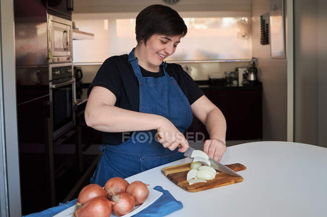 Веселая женщина в фартуке улыбается и режет сырой лук на разделочной доске на столе в кухне дома — стоковое фото