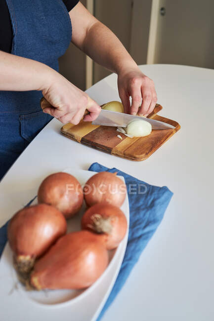 Женщина в фартуке режет сырой лук на разделочной доске на столе на кухне дома — стоковое фото