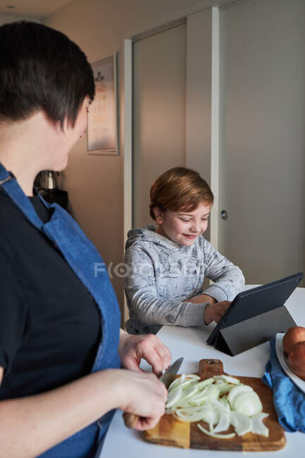 Rapaz feliz sorrindo e navegando tablet perto de mulher cortando cebola durante a preparação de alimentos na cozinha em casa — Fotografia de Stock