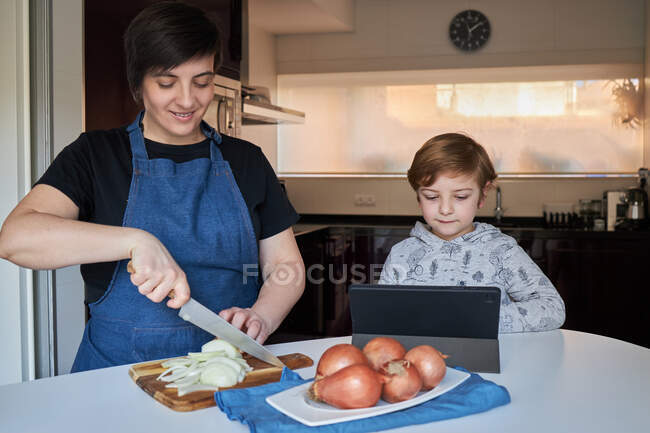 Мальчик улыбается и просматривает планшет рядом женщина режет лук во время приготовления пищи на кухне дома — стоковое фото