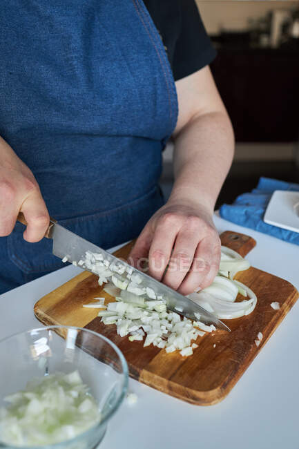 Mulher de colheita em avental cortando cebola crua na tábua de corte na mesa na cozinha em casa — Fotografia de Stock