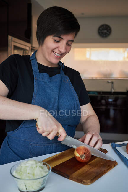 Fröhliche Frau in Schürze lächelt und schneidet rohe Zwiebeln auf Schneidebrett auf dem Tisch in der heimischen Küche — Stockfoto