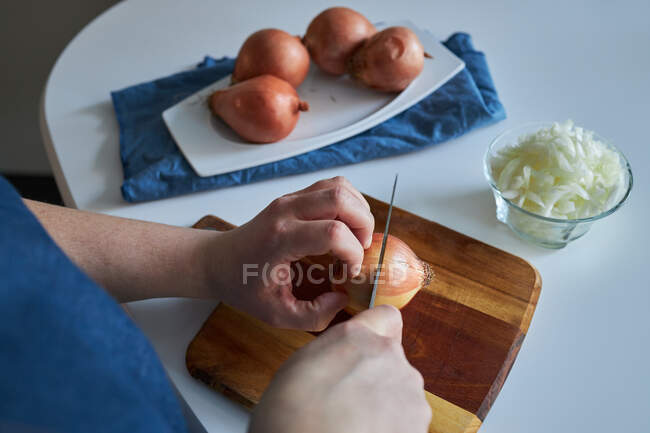 Erntefrau in Schürze schneidet rohe Zwiebel auf Schneidebrett auf dem Tisch in der heimischen Küche — Stockfoto