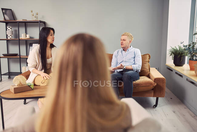 Pareja multiétnica sentada en el sofá y hablando de problemas mentales durante la sesión de terapia con el psicólogo - foto de stock