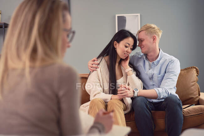 Conteúdo casal multirracial sentado no sofá no escritório do psicólogo e abraçando durante a consulta de psicoterapia — Fotografia de Stock
