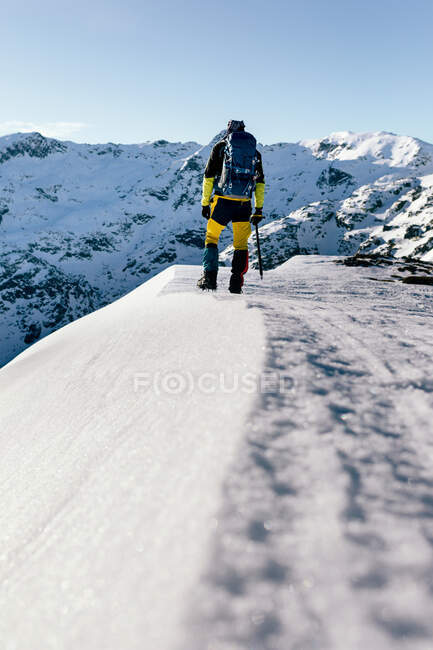 Полный вид назад на неузнаваемого альпиниста в теплой одежде с рюкзаком, стоящим на склоне заснеженной скалистой горы и наслаждающимся захватывающим пейзажем в солнечный зимний день — стоковое фото