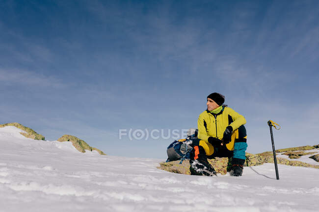 From below confident male mountaineer in stylish warm activewear with backpack an climbing equipment sitting on top of mountain and looking away while exploring snowy rocky landscape in sunny weather — Stock Photo