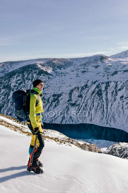 Full body side view of male mountaineer in warm activewear with backpack standing on slope of snowy rocky mountain and enjoying spectacular landscape in sunny winter day — Stock Photo