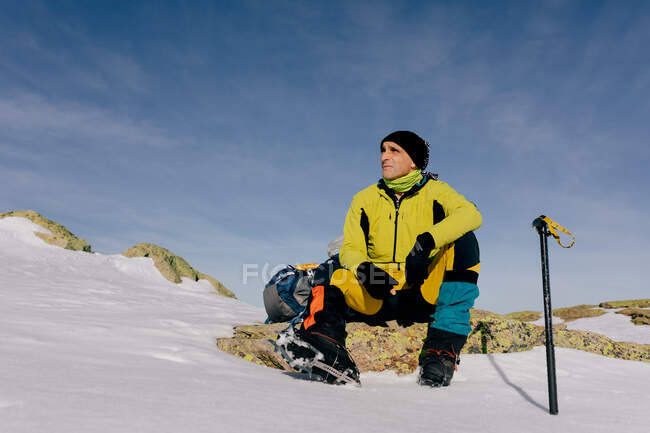 D'en bas alpiniste masculin confiant dans des vêtements de sport chauds élégants avec sac à dos un équipement d'escalade assis sur le sommet de la montagne et regardant loin tout en explorant un paysage rocheux enneigé par temps ensoleillé — Photo de stock