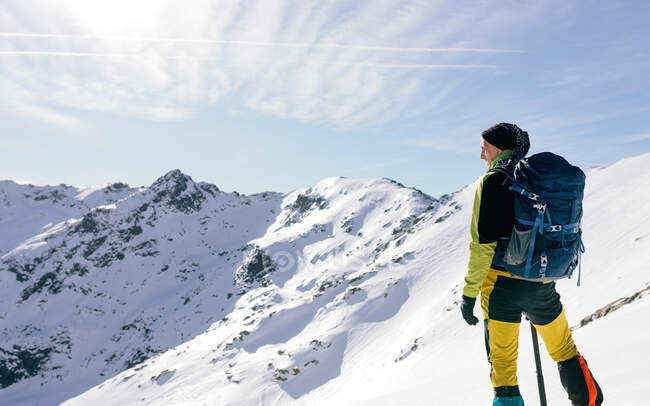 Back view of unrecognizable male mountaineer in warm activewear with backpack standing on slope of snowy rocky mountain and enjoying spectacular landscape in sunny winter day — Stock Photo