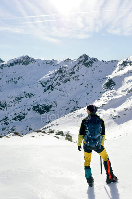 Full body back view of unrecognizable male mountaineer in warm activewear with backpack standing on slope of snowy rocky mountain and enjoying spectacular landscape in sunny winter day — Stock Photo