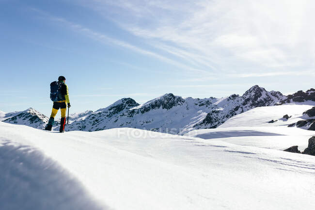 Полный вид назад на неузнаваемого альпиниста в теплой одежде с рюкзаком, стоящим на склоне заснеженной скалистой горы и наслаждающимся захватывающим пейзажем в солнечный зимний день — стоковое фото
