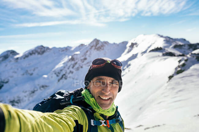 Alegre escalador masculino adulto en ropa deportiva cálida mirando a la cámara mientras toma selfie contra majestuosos picos rocosos nevados en un día soleado - foto de stock