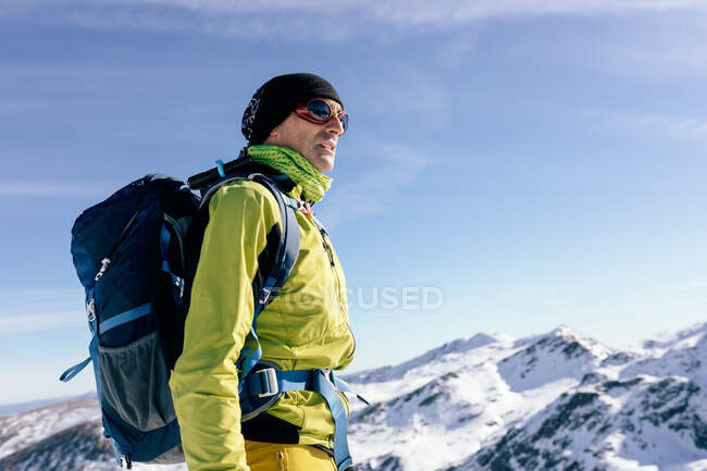 Снизу вид на самца альпиниста в теплой одежде с рюкзаком, стоящим на склоне заснеженной скалистой горы и наслаждающимся захватывающим пейзажем в солнечный зимний день — стоковое фото