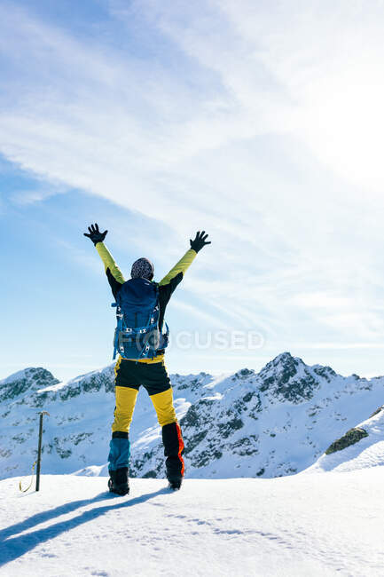 Full body back view of unrecognizable active male mountaineer in colorful activewear raising arms and enjoying freedom while standing on snowy top of mountain against blue cloudy sky in sunny day in highlands — Stock Photo