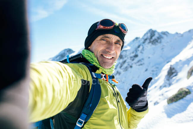 Fröhlicher erwachsener männlicher Bergsteiger in warmer Aktivkleidung blickt in die Kamera, während er bei sonnigem Wetter ein Selfie mit majestätischen schneebedeckten Felsgipfeln macht — Stockfoto