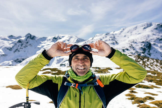 Confident happy male mountaineer in stylish warm activewear with backpack an climbing equipment standing on top of mountain and looking at camera while exploring snowy rocky landscape in sunny weather — Stock Photo