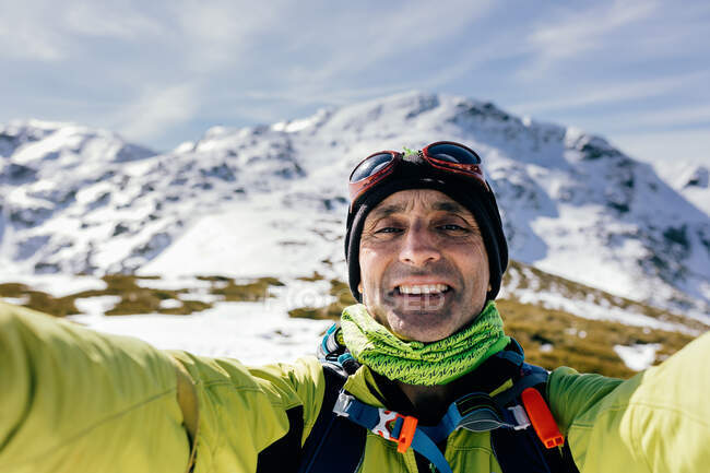 Веселый взрослый альпинист в теплой одежде смотрит в камеру, делая селфи против величественных снежных скалистых вершин в солнечный день — стоковое фото