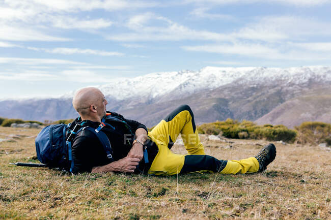 Vista laterale completa del corpo dell'alpinista maschio attivo adulto in activewear con zaino che si riposa sulla cima della montagna erbosa e gode di una pittoresca vista della gamma coperta di neve — Foto stock