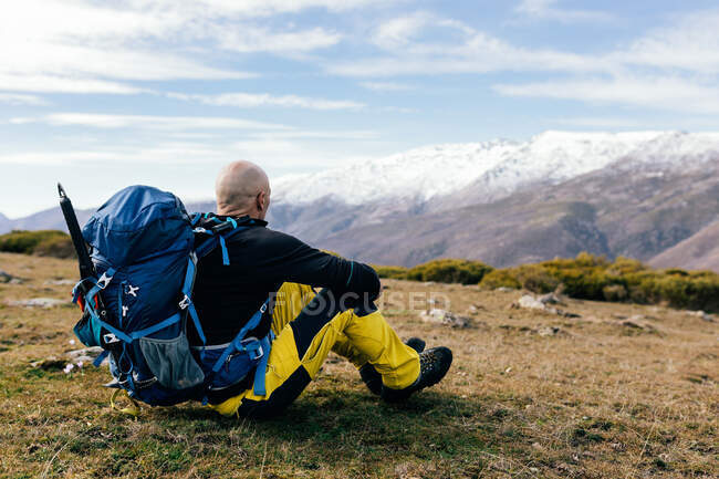 Full body side view of adult active male mountaineer in activewear with backpack getting rest on grassy mountain top and enjoying picturesque view of snow covered range — Stock Photo